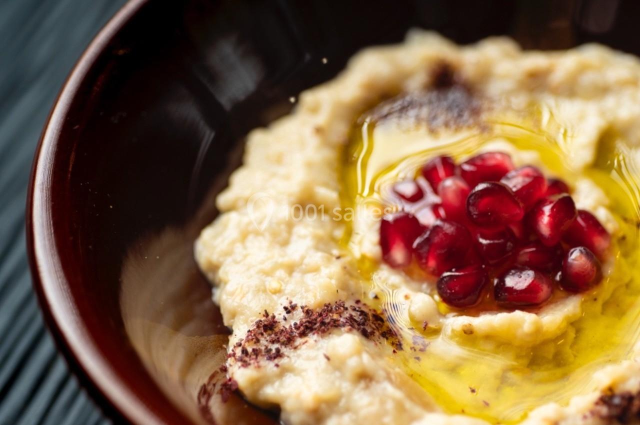 Bol de houmous garni de graines de grenade, d'huile d'olive et de sumac, posé sur une surface noire texturée.