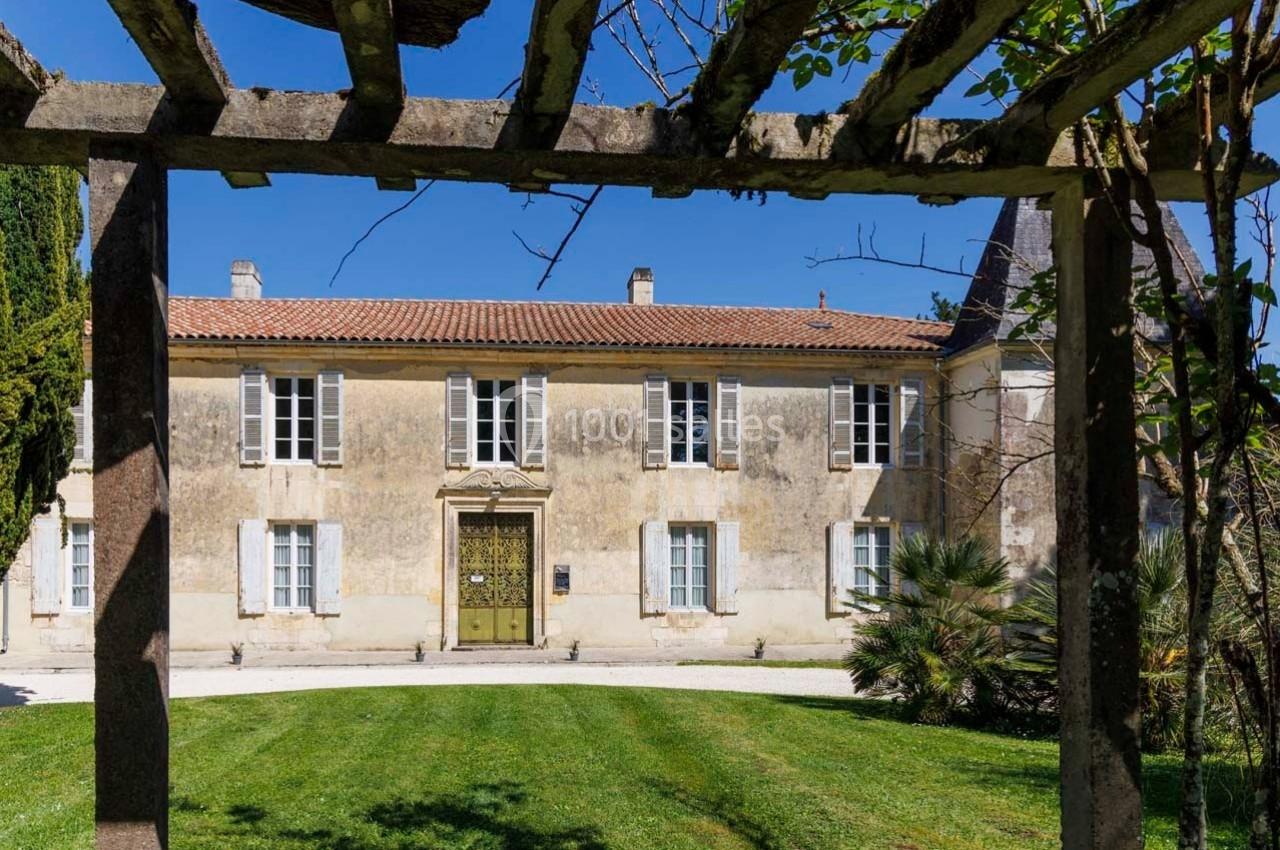 Façade d'une maison ancienne en pierre avec toit en tuiles, vue à travers une pergola en bois, entourée de verdure.