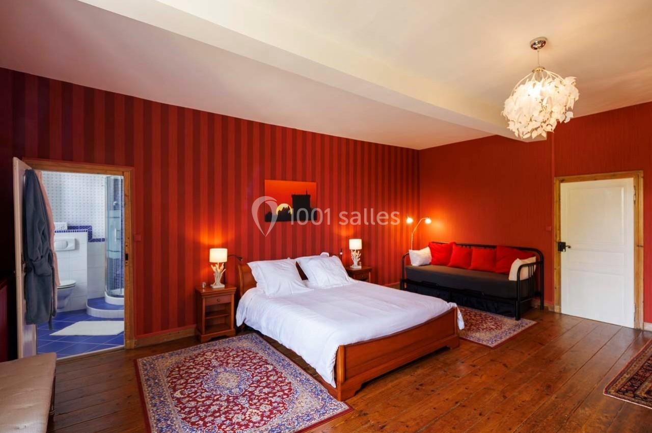 Chambre avec lit double, canapé, murs rouges, parquet, tapis, et salle de bain attenante avec carrelage bleu.
