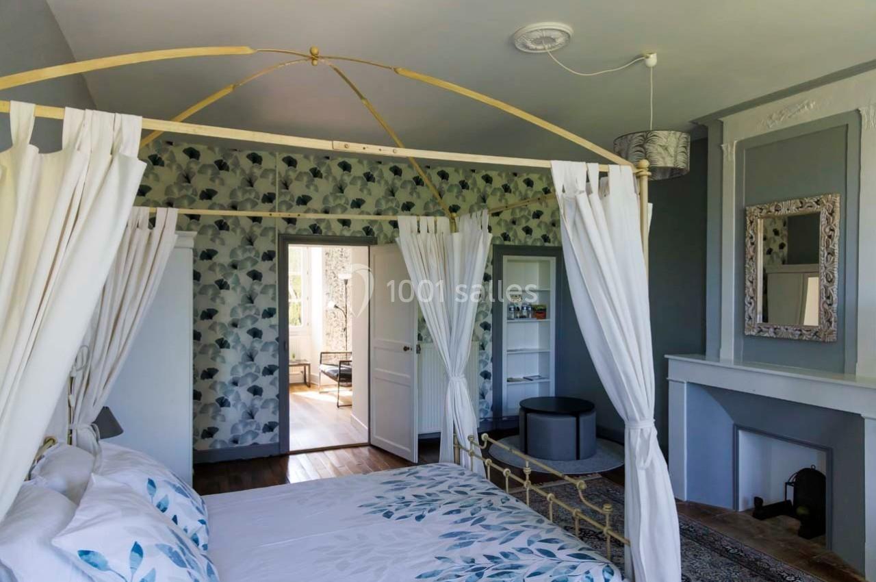 Chambre lumineuse avec lit à baldaquin, murs décorés de papier peint floral et cheminée ornée d'un miroir.