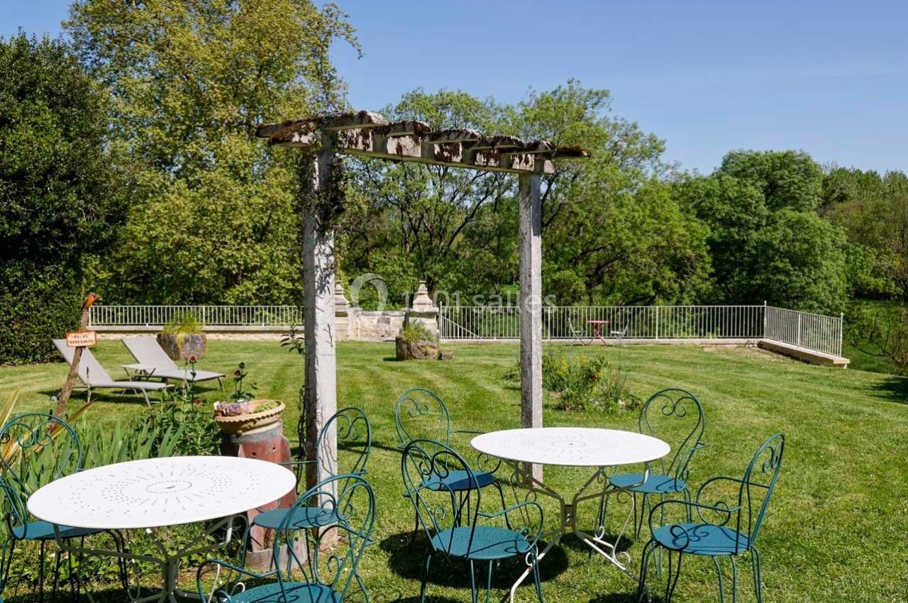 Tables et chaises en métal sur une pelouse, entourées de verdure avec une pergola en bois et des transats à l'arrière.