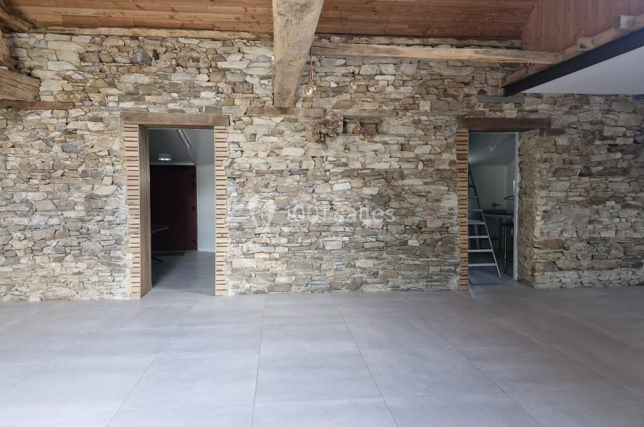 Mur en pierre apparente avec deux ouvertures donnant sur des pièces adjacentes, plafond en bois et sol carrelé.