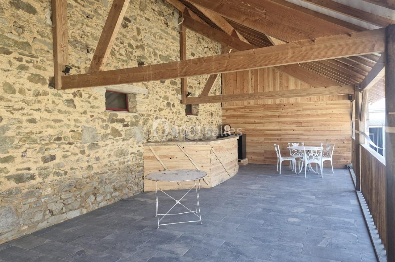 Terrasse couverte avec murs en pierre, cuisine extérieure en bois, table ronde et chaises blanches sur sol en carrelage gris.