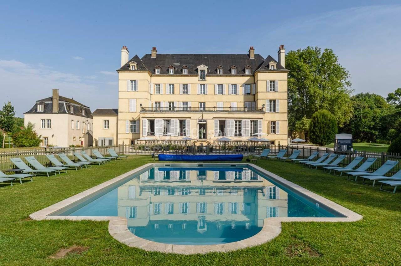 Façade d'un château entouré de verdure avec une piscine rectangulaire et des transats alignés au premier plan.