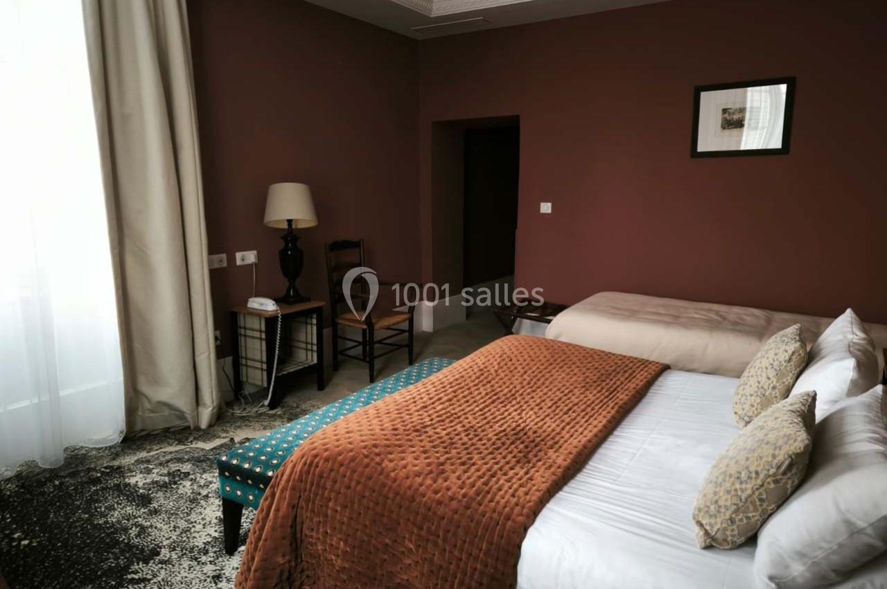 Chambre avec murs rouges, lit double, lit simple, fauteuil, lampe, rideaux clairs et tapis sombre.