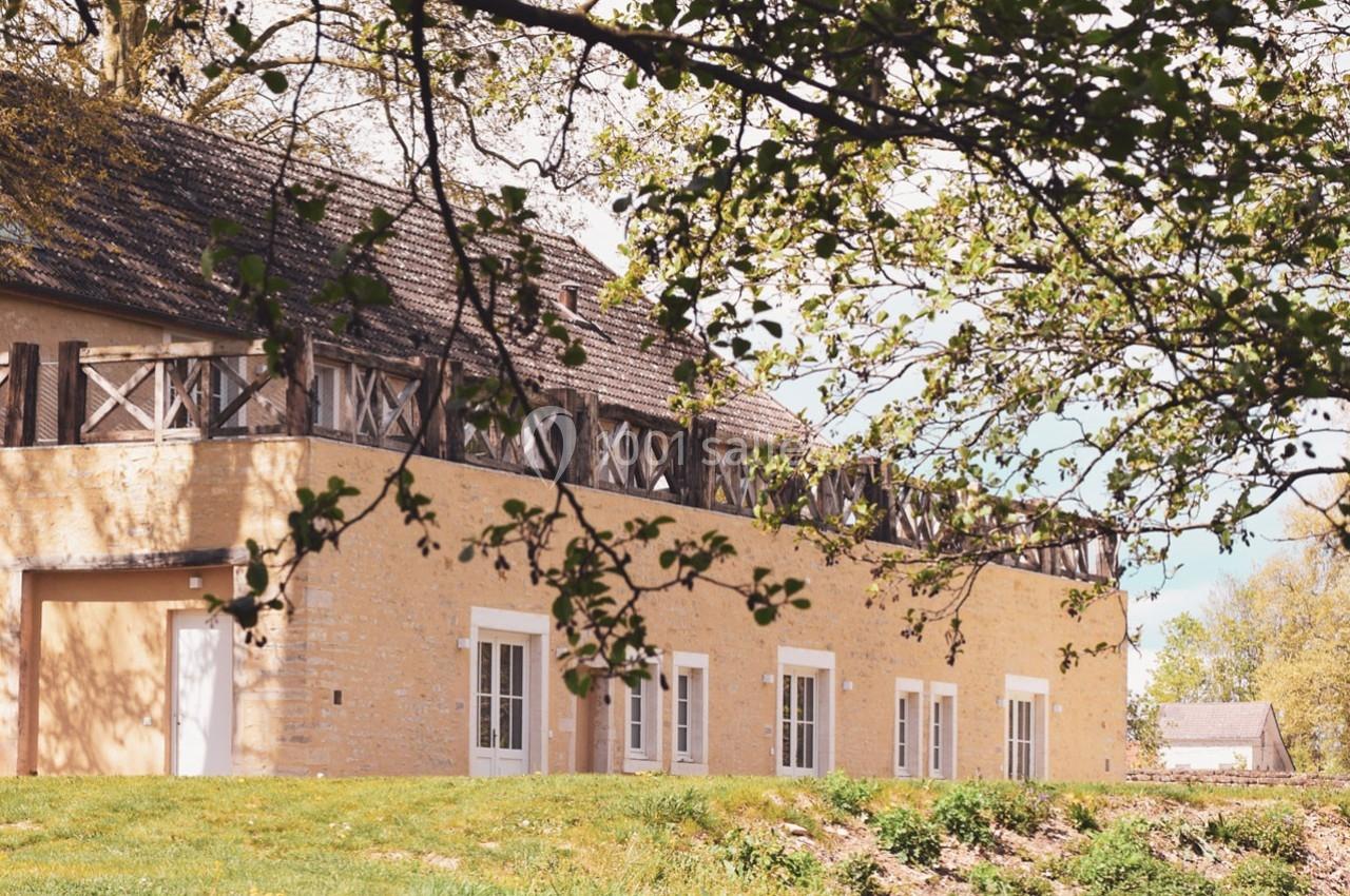 Bâtiment en pierre beige avec des fenêtres blanches, entouré de verdure et partiellement ombragé par des branches d'arbres.