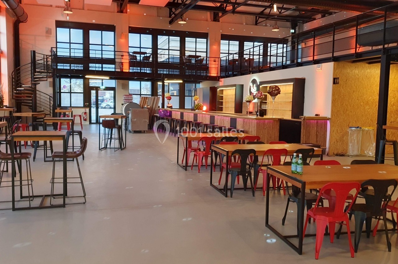 Salle moderne avec tables en bois, chaises colorées et bar éclairé, dans un espace lumineux à mezzanine.