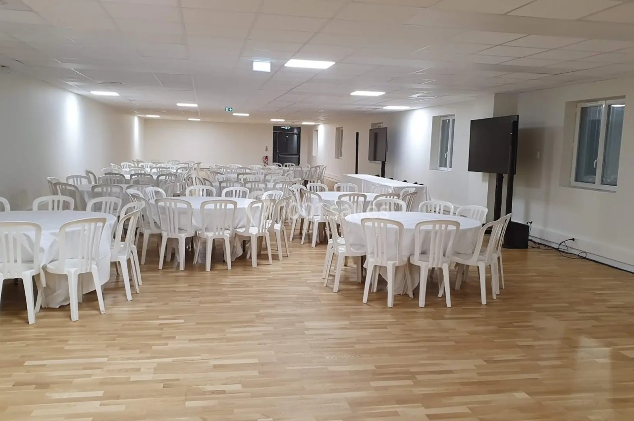 Salle vide avec parquet, tables rondes blanches et chaises en plastique alignées, éclairée par des néons au plafond.