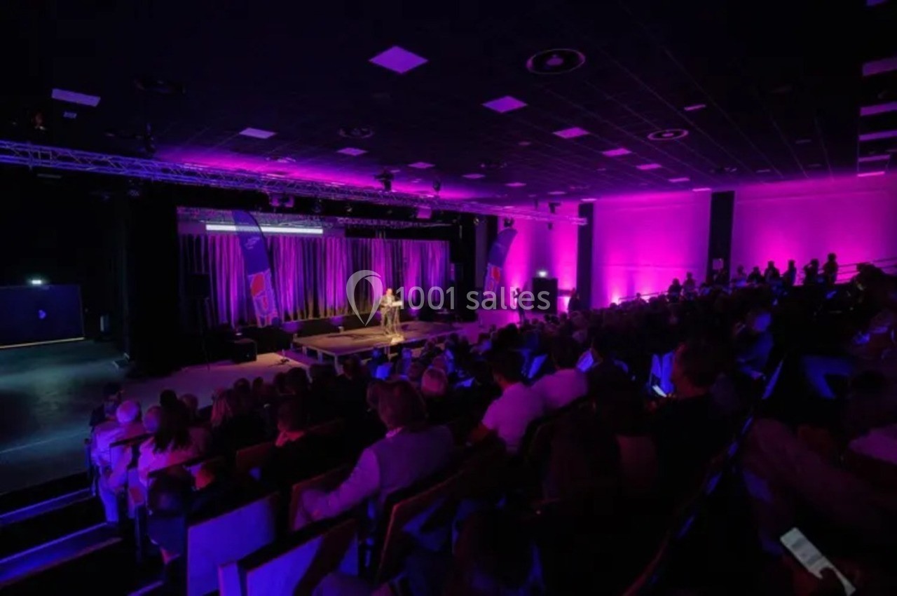 Salle de conférence éclairée en violet, avec un orateur sur scène et un public assis dans l'obscurité.