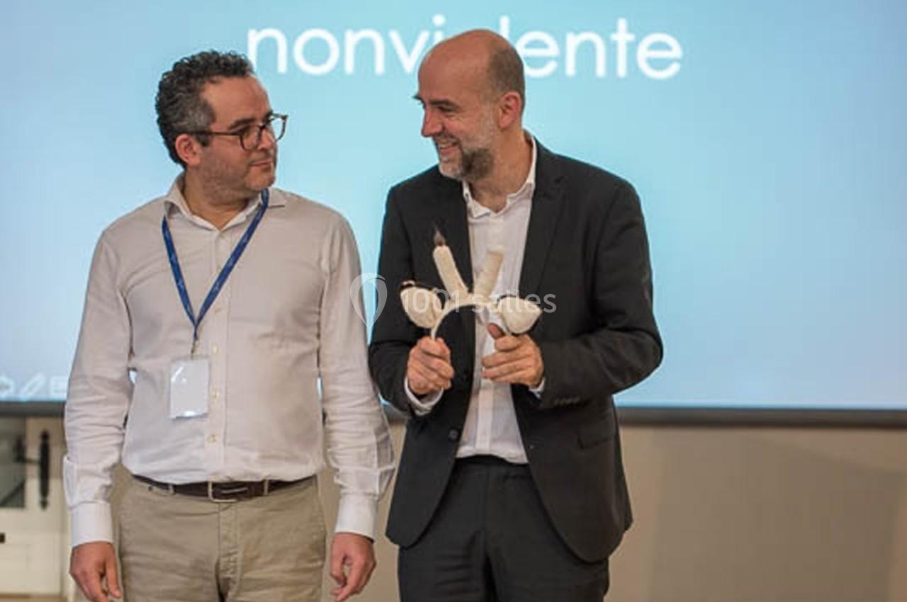 Deux hommes debout, l'un tenant un objet en forme de vertèbre, devant un écran avec le mot ’nonvidente’.