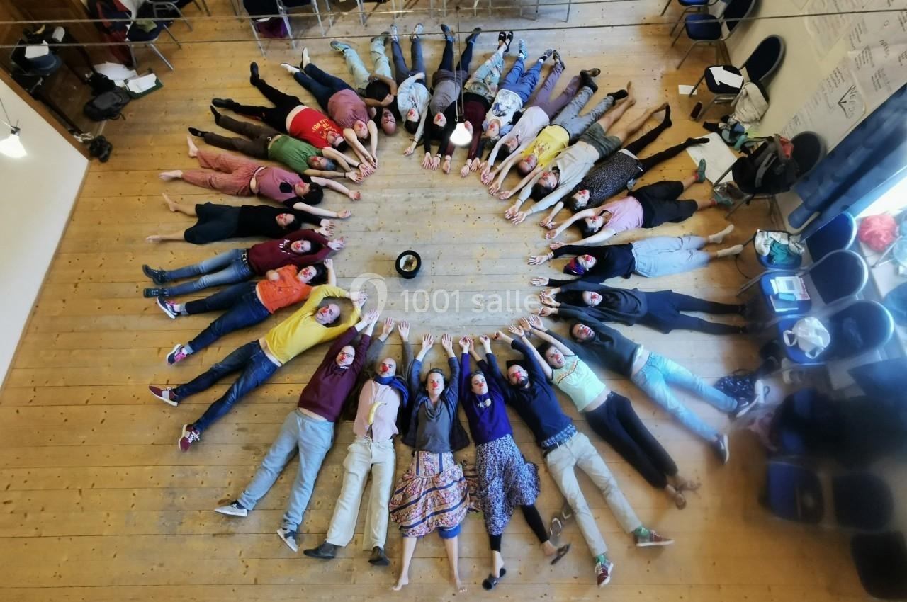 Des personnes allongées en cercle sur un sol en bois, bras tendus vers le centre où se trouve un objet noir.