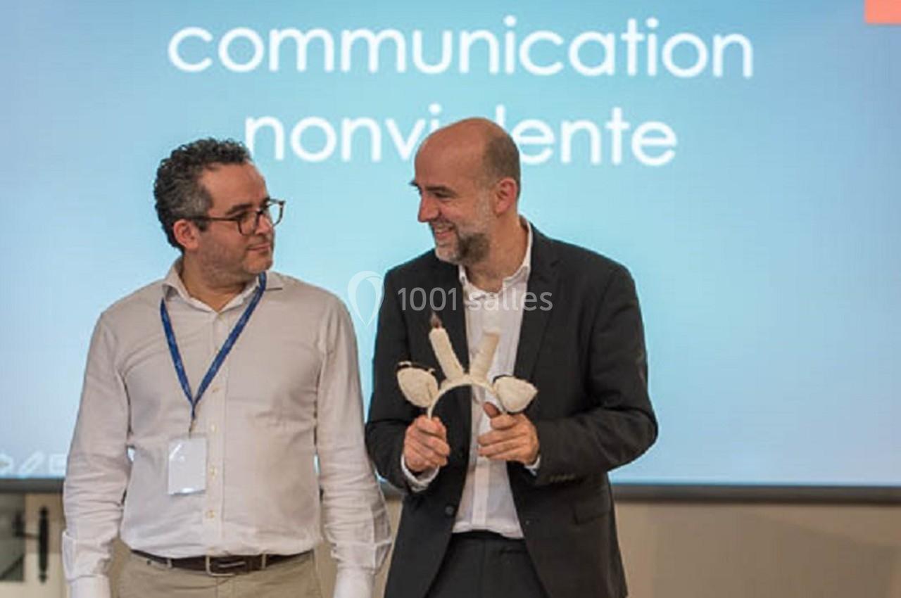 Deux hommes discutent devant un écran affichant ’communication non violente’, l'un tenant un objet en bois.