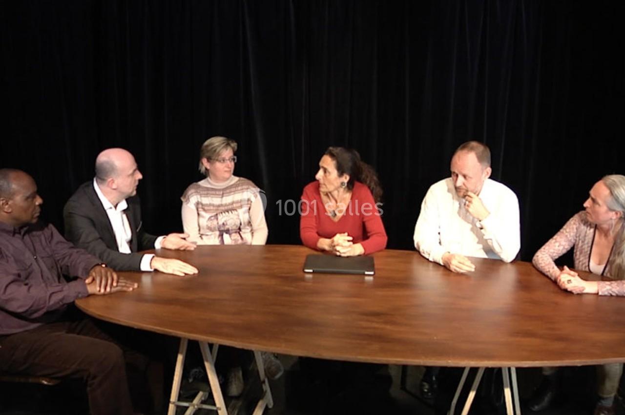 Six personnes discutent autour d'une table ovale en bois, devant un rideau noir.