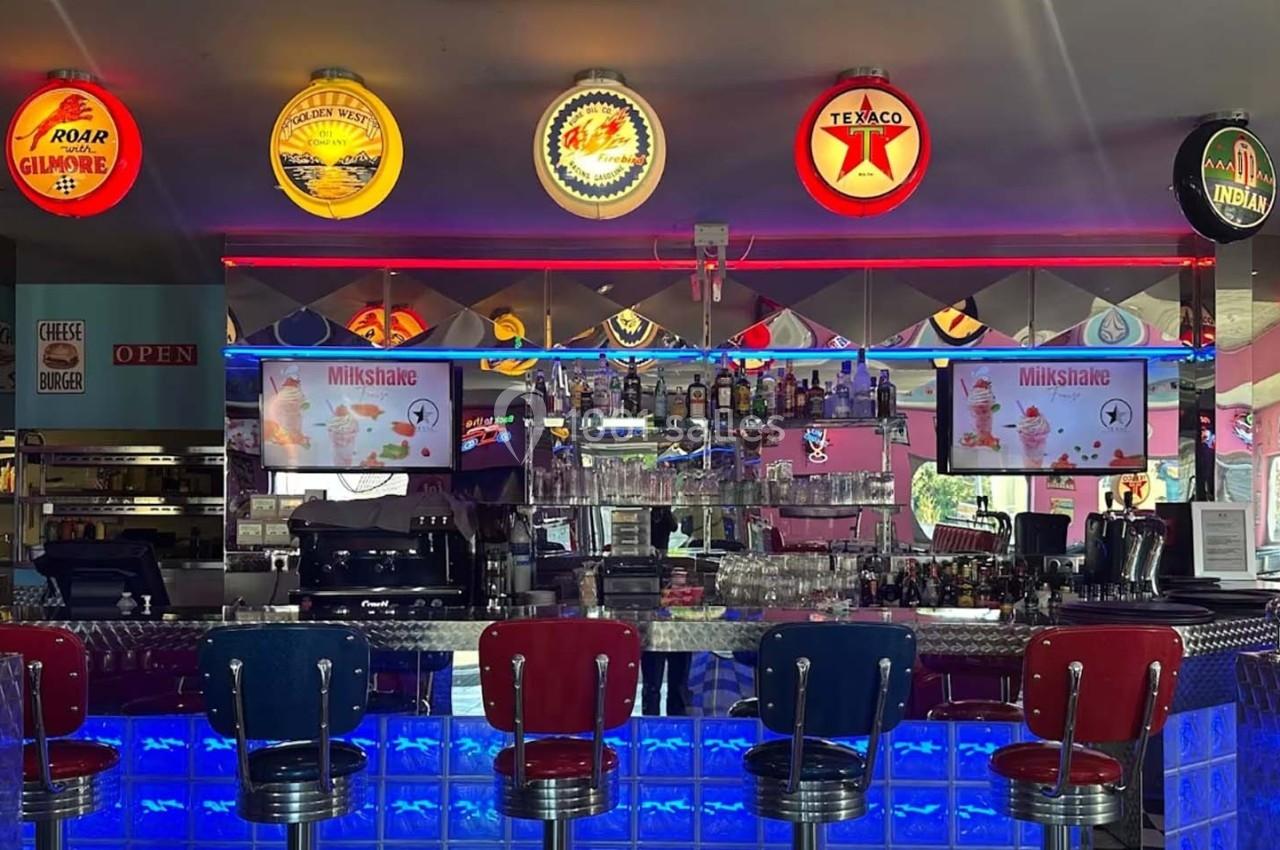Intérieur d'un diner rétro avec un bar éclairé en bleu, tabourets rouges et enseignes lumineuses vintage au mur.