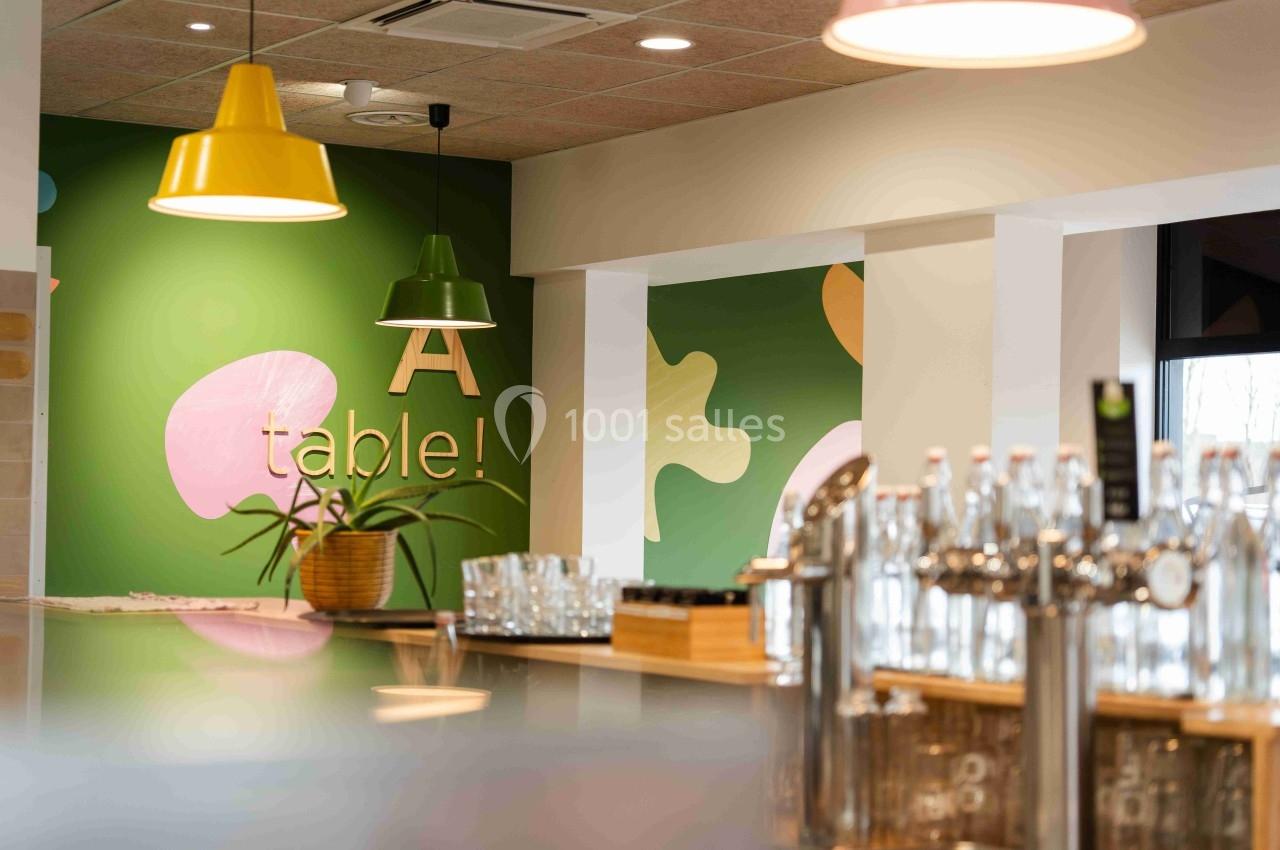 Intérieur d'un café moderne avec comptoir, plantes, luminaires colorés et mur décoré d'éléments graphiques.