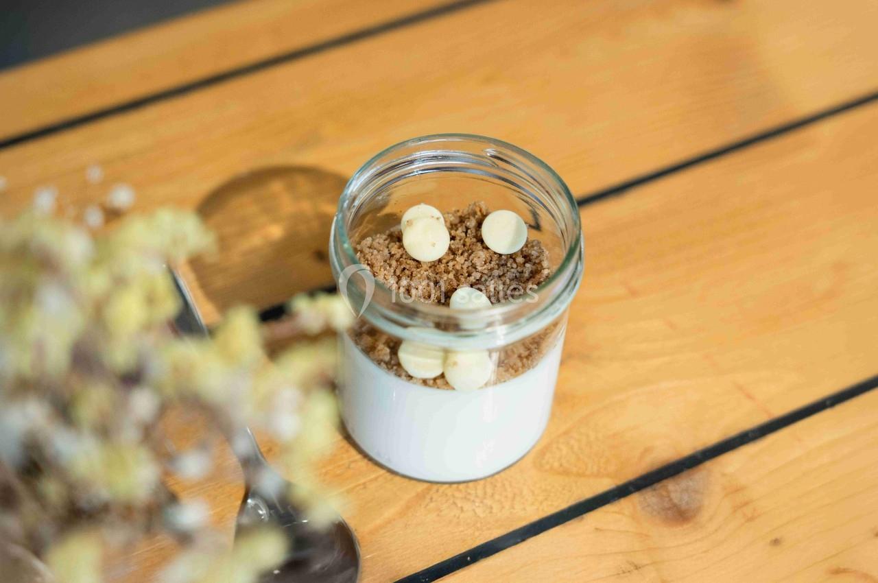 Dessert en pot composé de crème blanche, crumble et pépites de chocolat blanc, posé sur une table en bois.