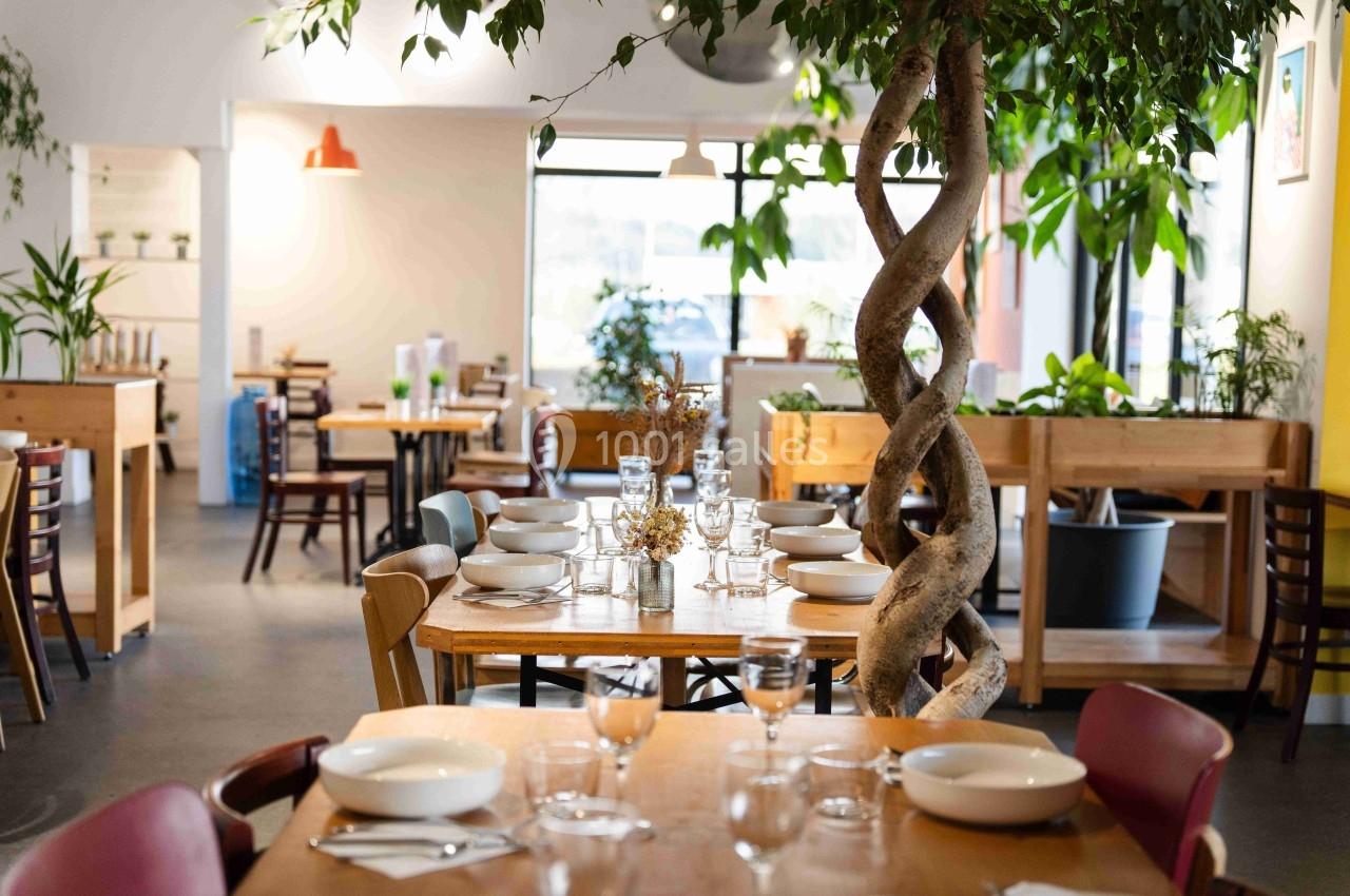 Salle de restaurant lumineuse avec des tables dressées, des plantes vertes et un arbre décoratif au centre.