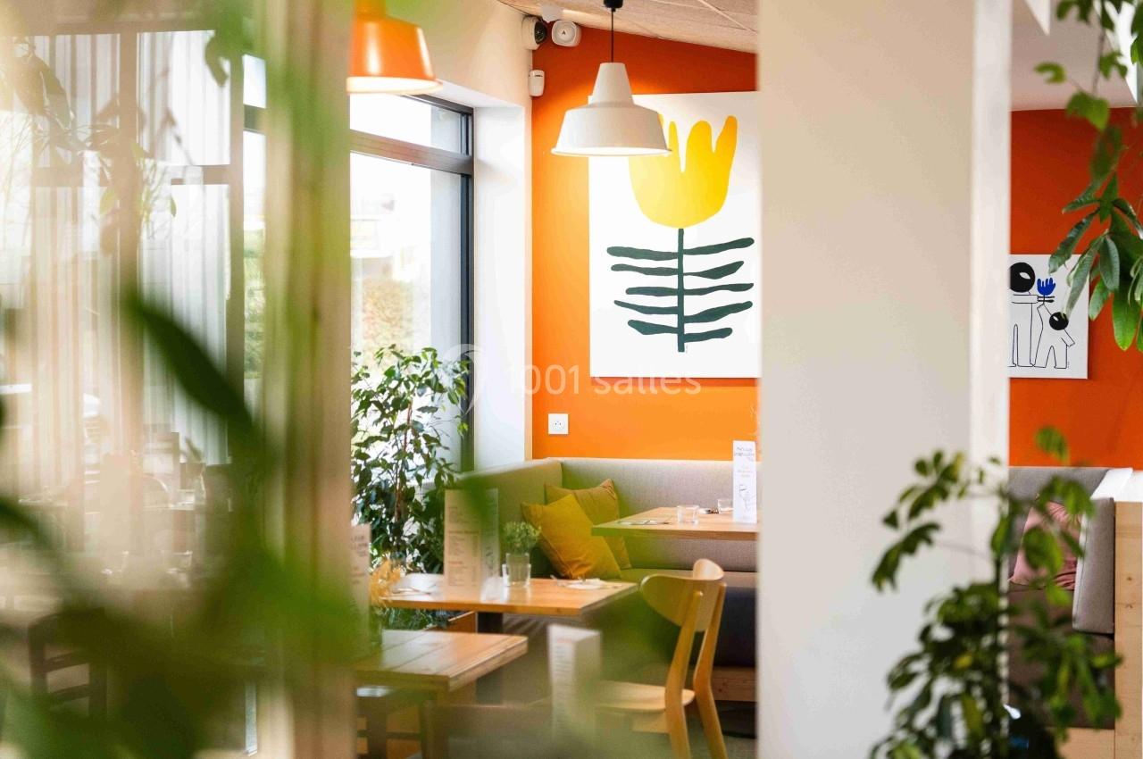 Intérieur lumineux d’un café moderne avec des plantes, des tables en bois et un mur orange décoré d’une peinture florale.