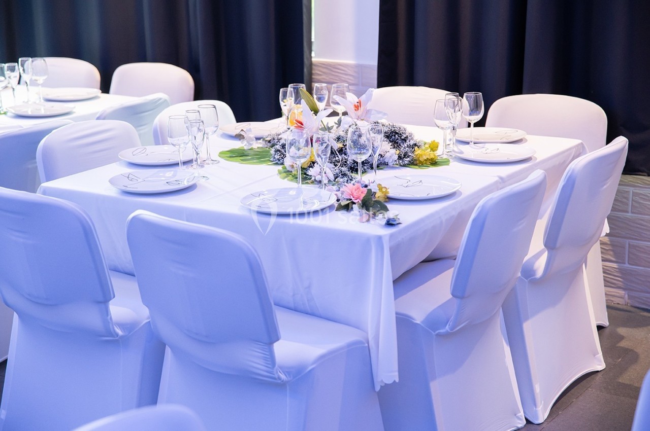 Table dressée avec nappes blanches, vaisselle, verres et centre de table floral dans une salle élégante.