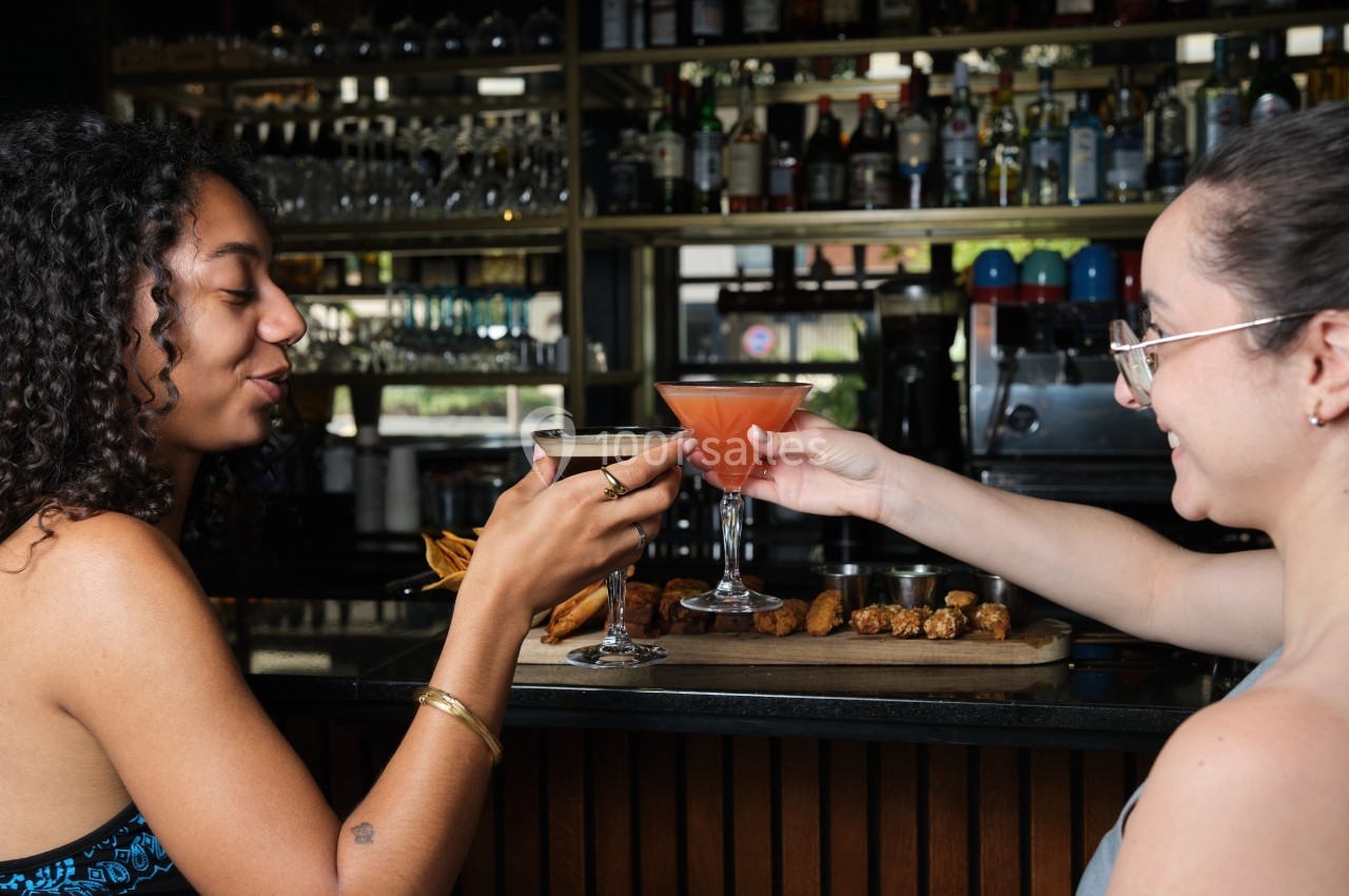 Deux femmes trinquent avec des cocktails devant un bar, des amuse-bouches posés sur le comptoir.