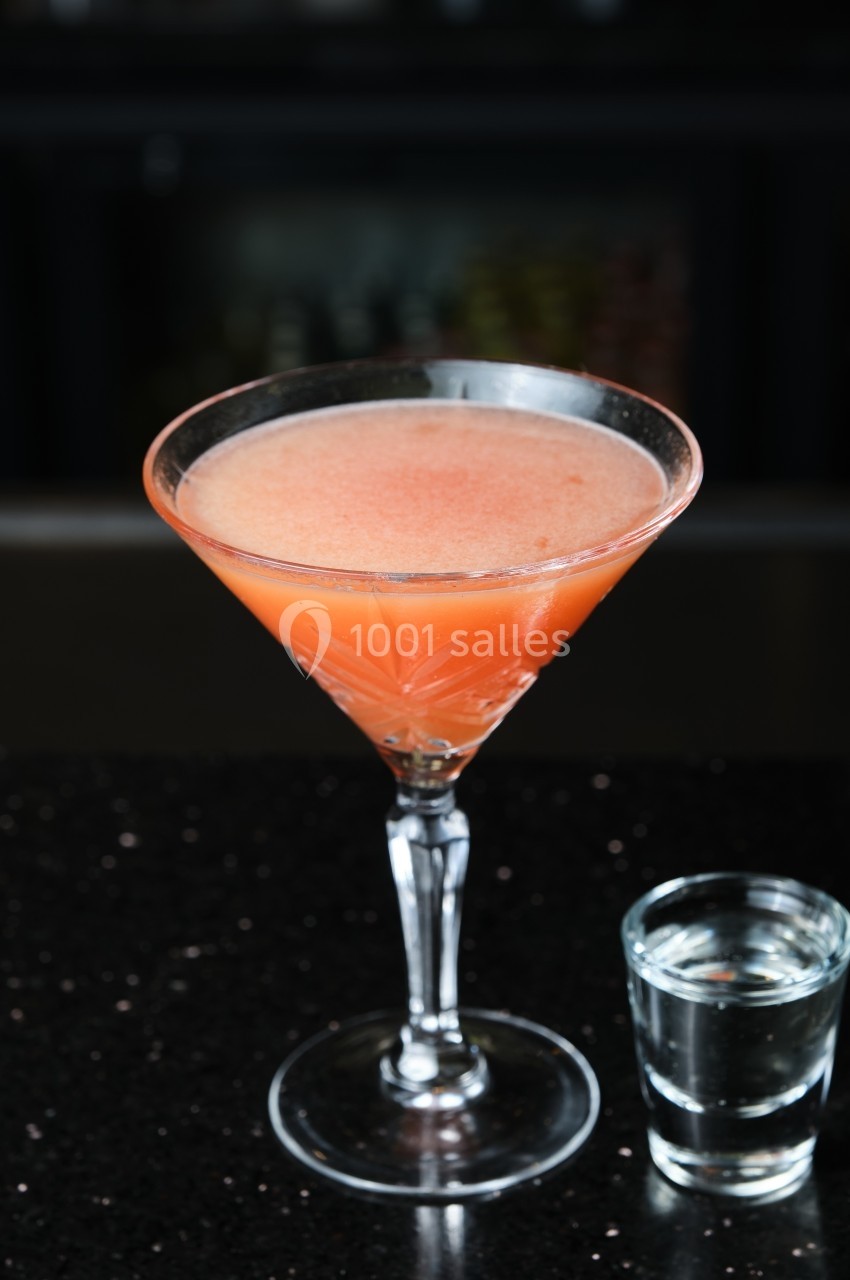 Verre à cocktail orange sur une table noire, accompagné d'un petit verre d'eau transparente.