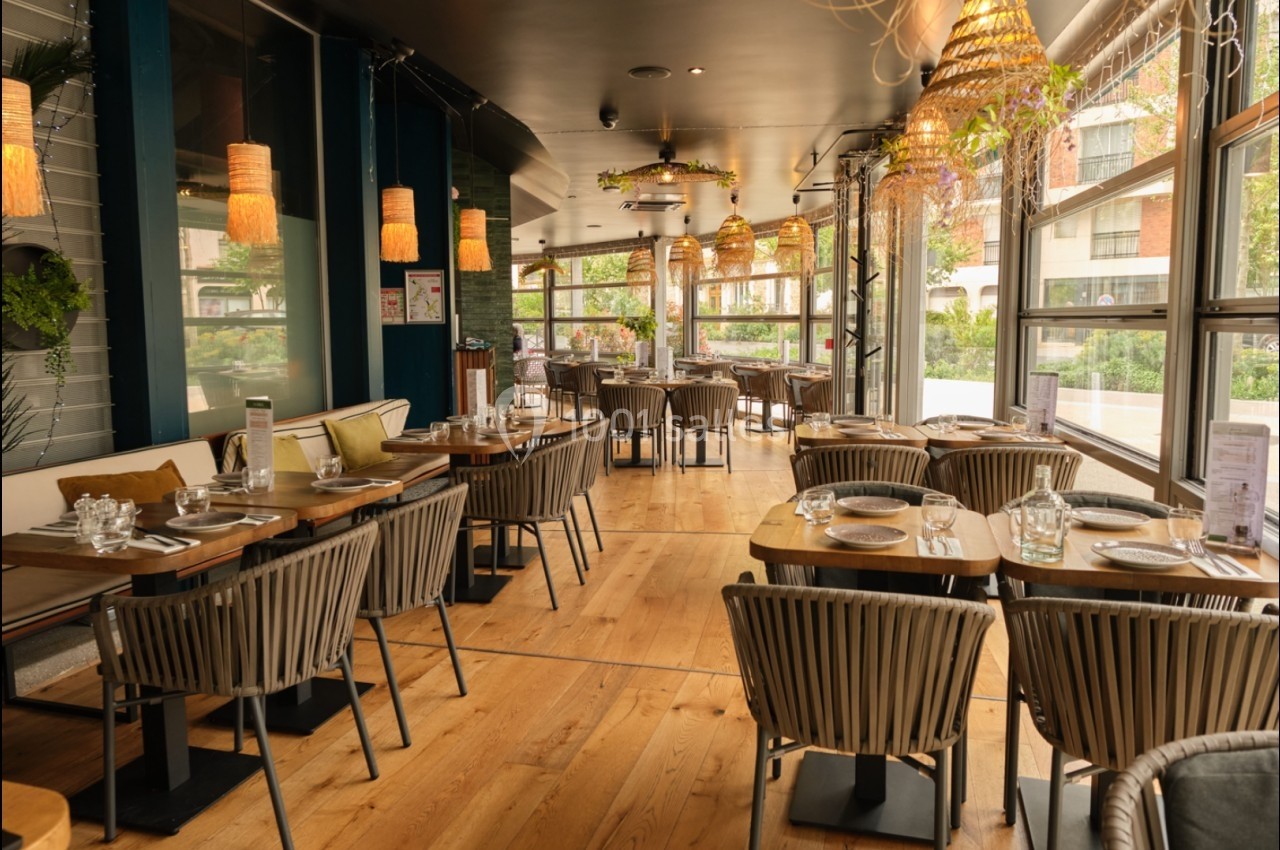 Salle de restaurant lumineuse avec tables dressées, chaises en rotin et grandes baies vitrées donnant sur un espace…