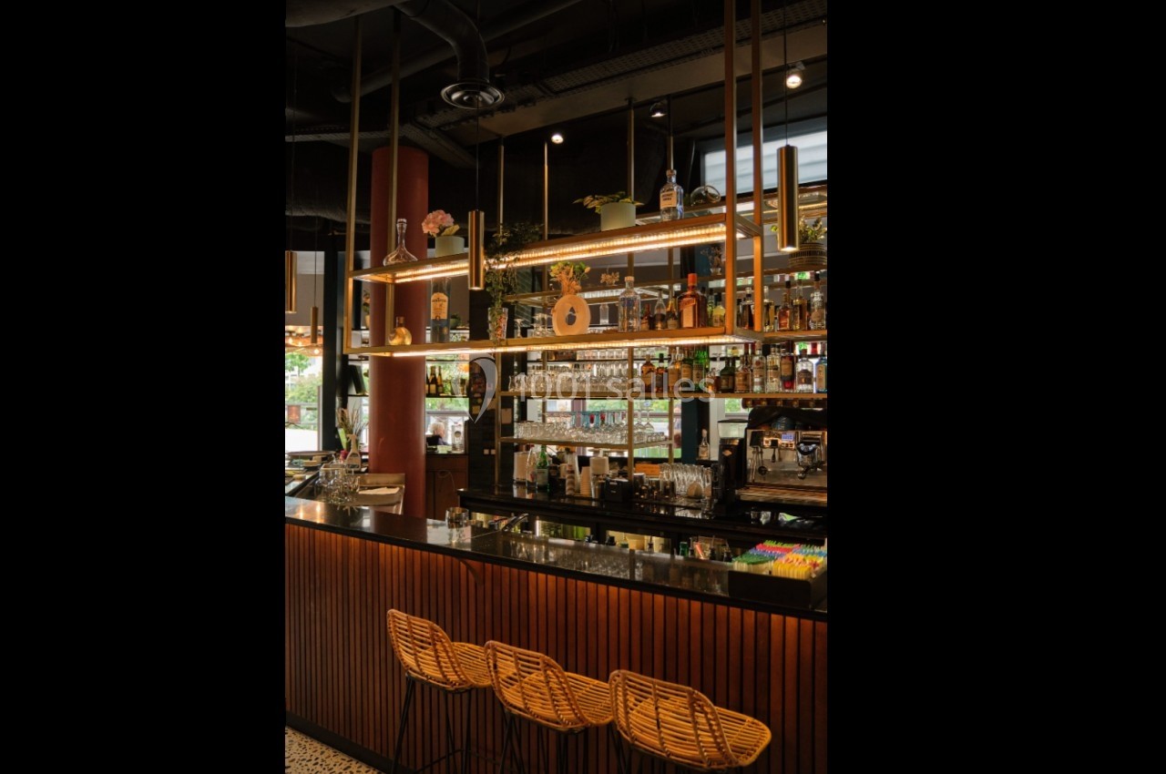Bar moderne avec comptoir en bois, tabourets en rotin, étagères éclairées et divers alcools exposés.