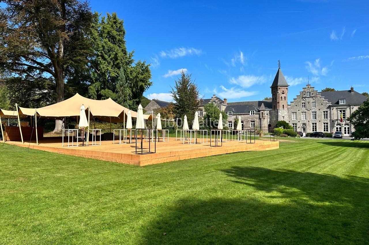 Terrasse en bois avec parasols et tentes, installée sur une pelouse devant un bâtiment historique sous un ciel bleu.