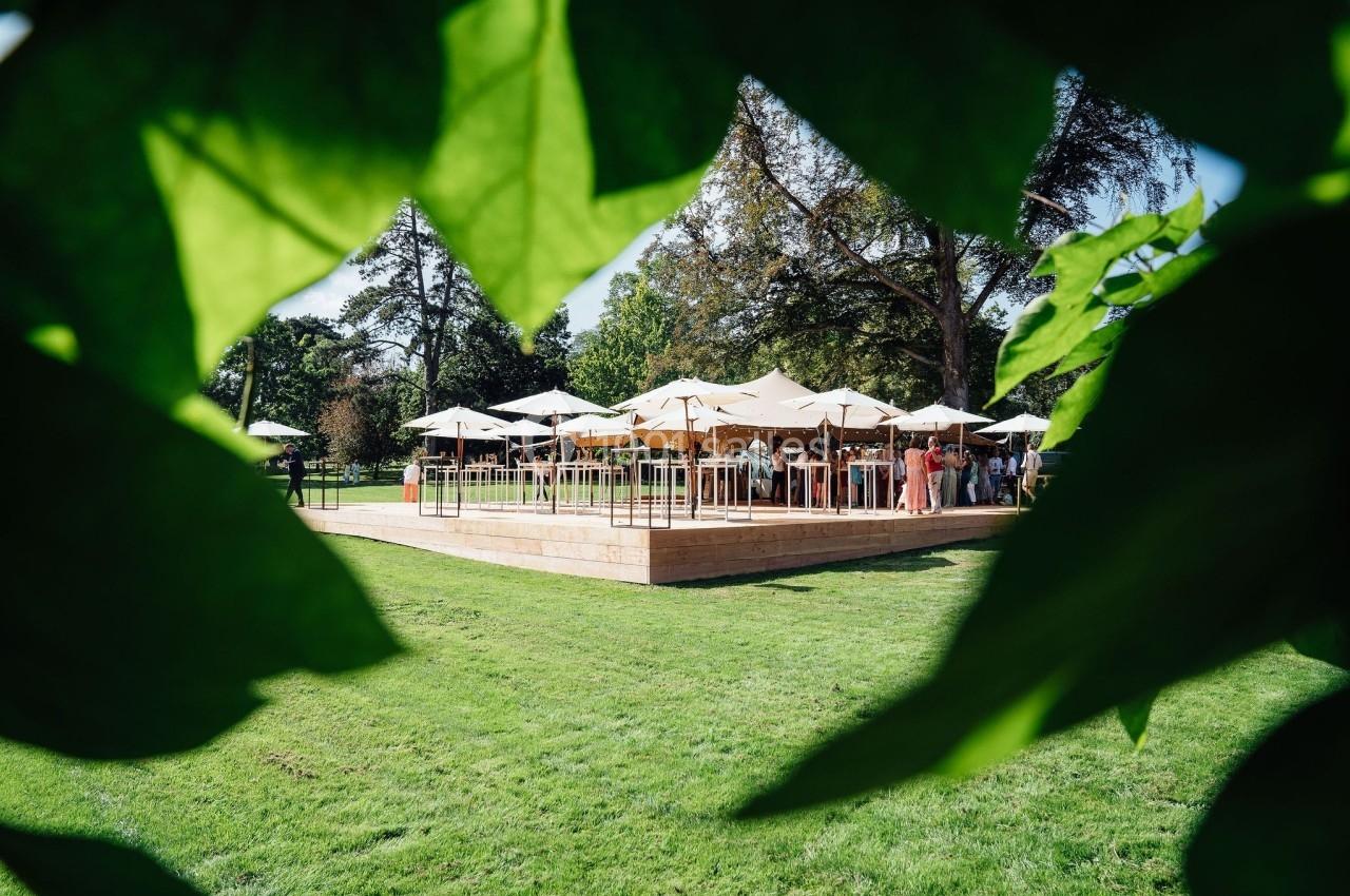 Terrasse en bois avec parasols blancs dans un parc verdoyant, vue à travers des feuilles au premier plan.