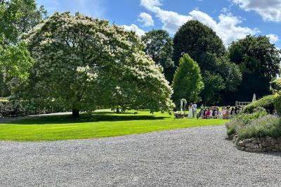 Miniature Location salle Namur (Namur) - Château de Beez #18 Groupe de personnes assises en extérieur lors d'une cérémonie, entourées d'arbres et de fleurs sous un ciel partiellement…