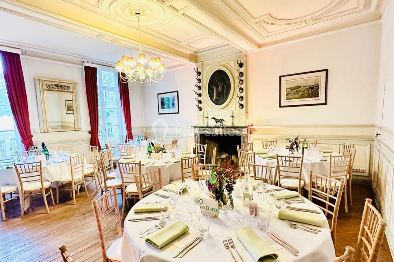 Salle élégante avec tables rondes dressées pour un repas, décorée de fleurs, avec moulures au plafond et cheminée.