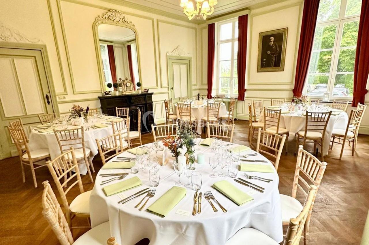 Salle élégante avec tables rondes dressées, nappes blanches, chaises en bois clair et grandes fenêtres laissant entrer la…