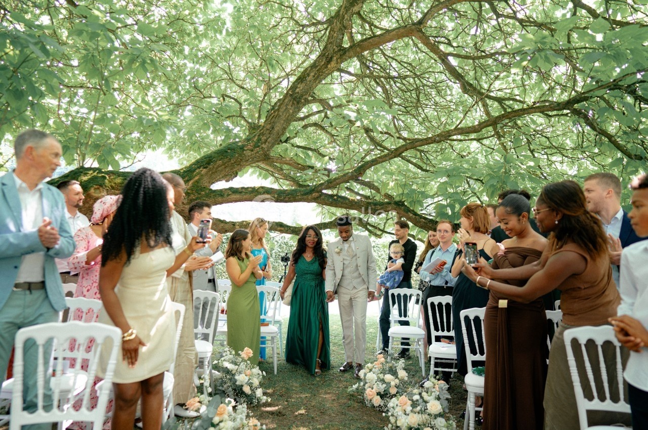 Un couple marche dans une allée fleurie sous un grand arbre, entouré d'invités applaudissant et prenant des photos.