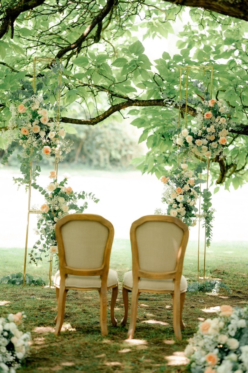 Deux chaises en bois devant une arche florale décorée de roses et feuillages, sous un arbre dans un jardin.