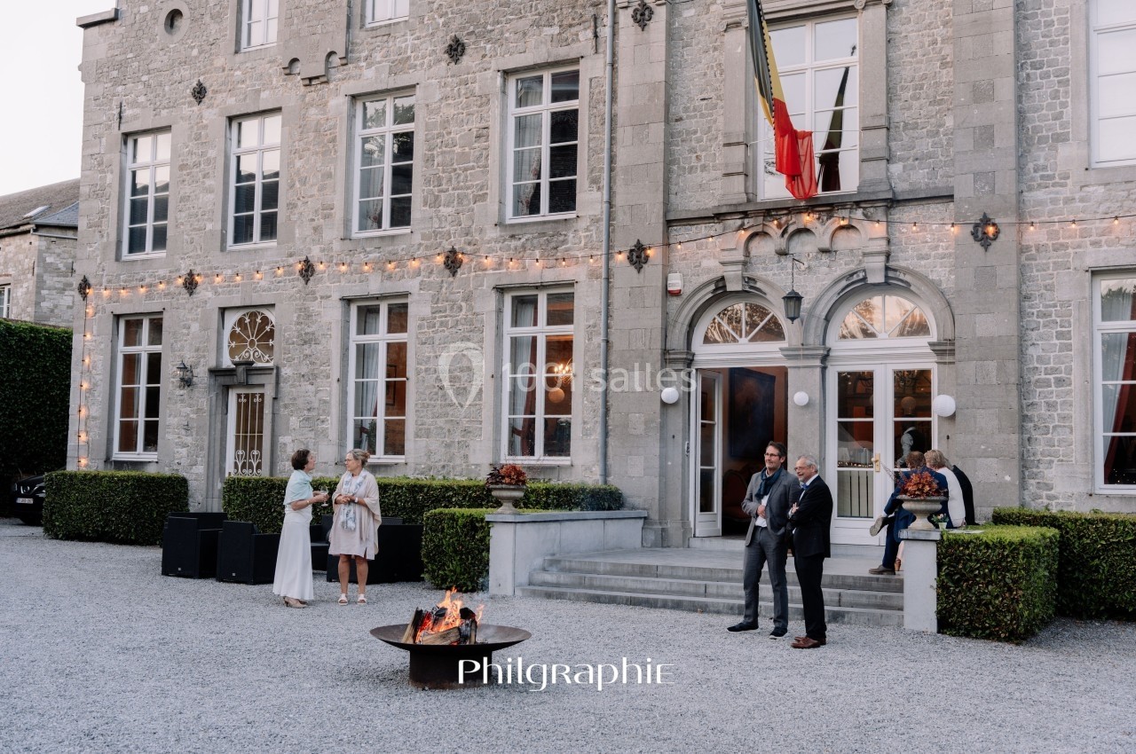 Façade d'un bâtiment en pierre avec des invités discutant près d'un brasero allumé dans une cour gravillonnée.