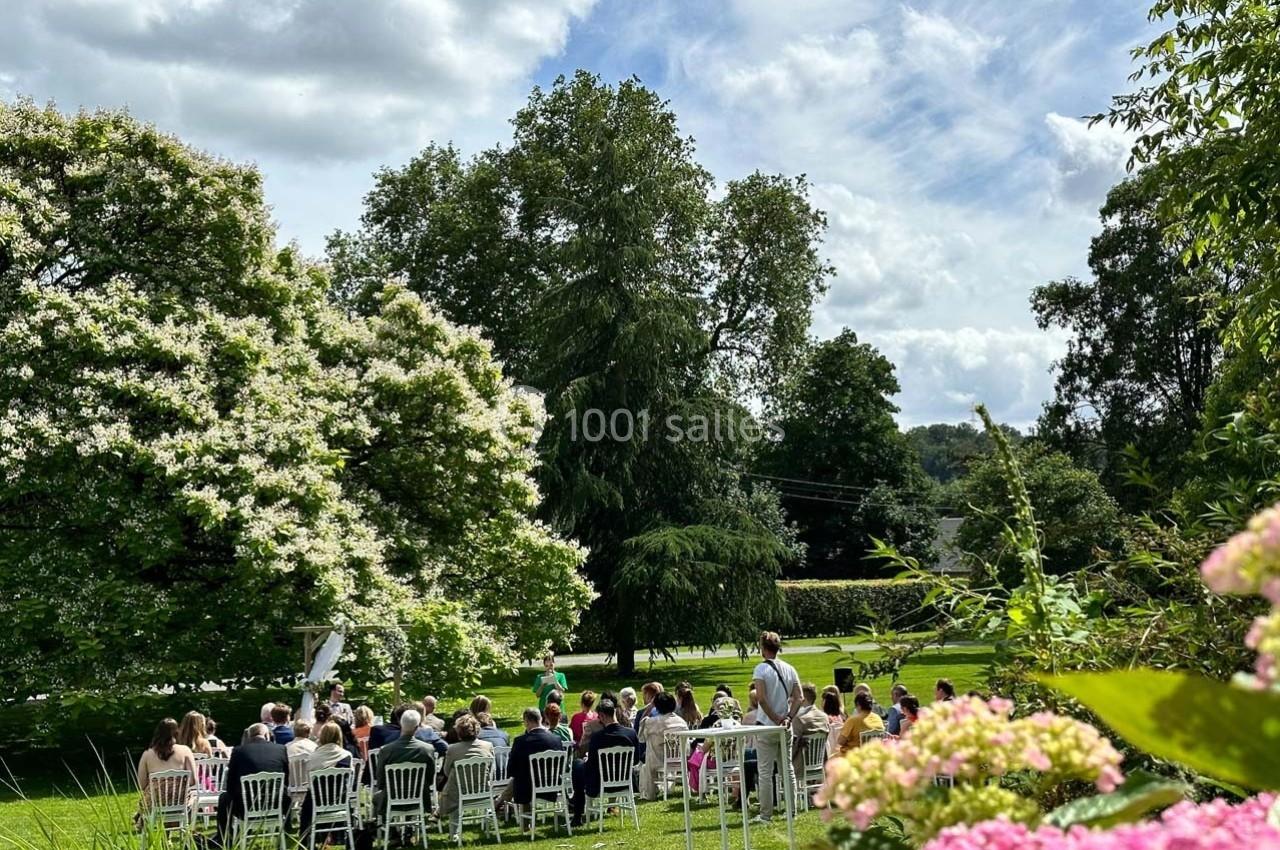 Groupe de personnes assises en extérieur lors d'une cérémonie, entourées d'arbres et de fleurs sous un ciel partiellement…