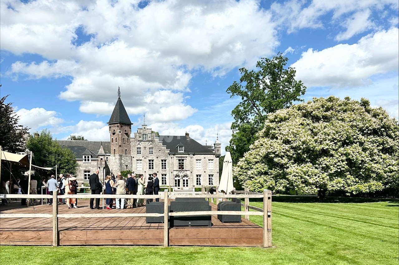 Groupe de personnes rassemblées sur une terrasse en bois devant un château entouré de pelouses et d'arbres sous un ciel…