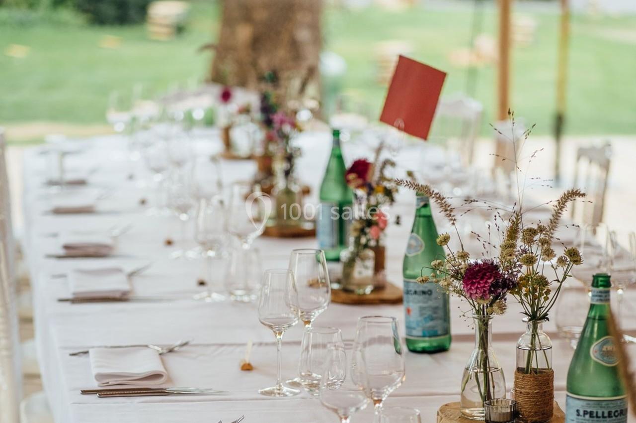 Table dressée en extérieur avec nappes blanches, verres, bouteilles d'eau et bouquets de fleurs champêtres.