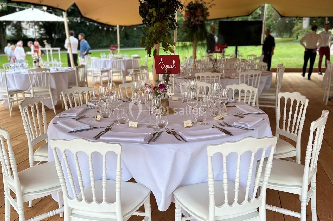 Table ronde élégamment dressée avec nappes blanches, chaises assorties et décoration florale sous une tente en extérieur.