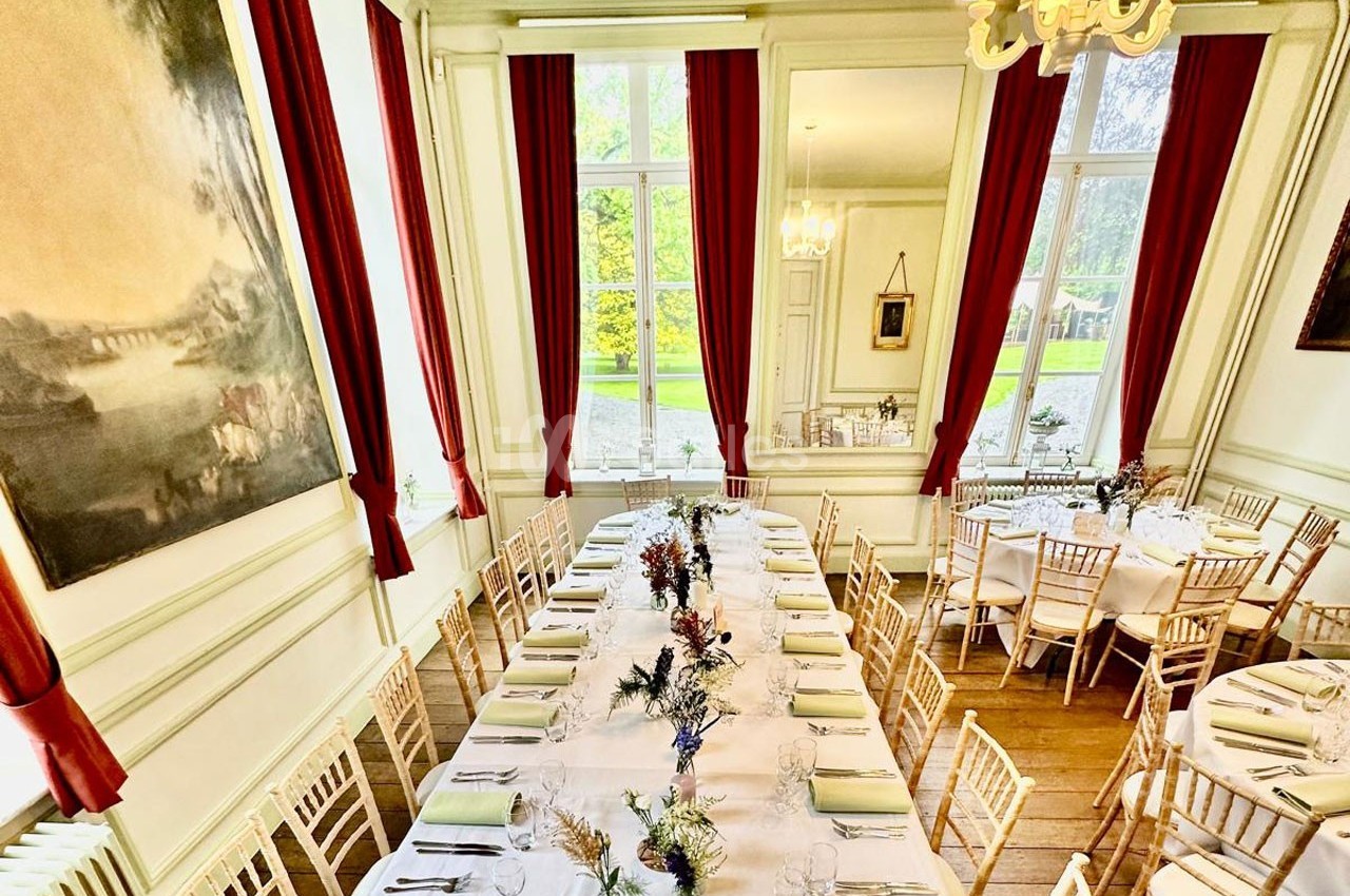 Salle lumineuse avec de grandes fenêtres, tables dressées pour un repas, chaises en bois clair et rideaux rouges.