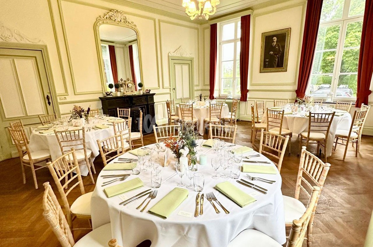 Salle élégante avec tables rondes dressées, nappes blanches, chaises en bois clair et grandes fenêtres laissant entrer la…