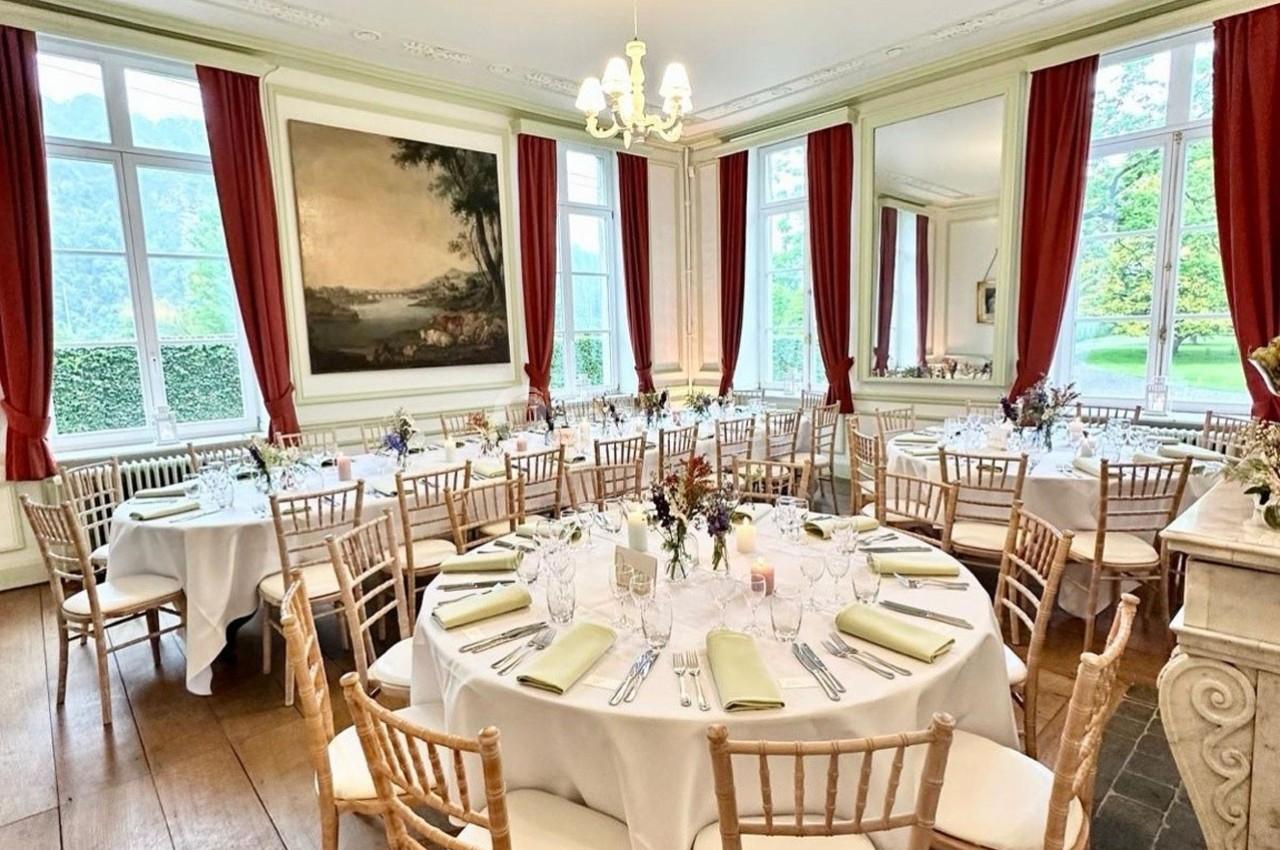 Salle élégante avec tables rondes dressées pour un repas, chaises en bois, grandes fenêtres et rideaux rouges.