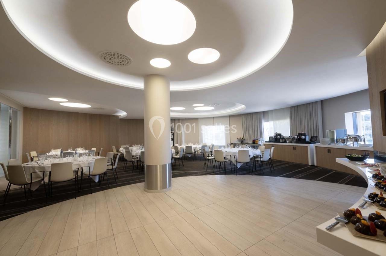 Salle de réception lumineuse avec tables dressées, buffet en arrière-plan et éclairage moderne au plafond.