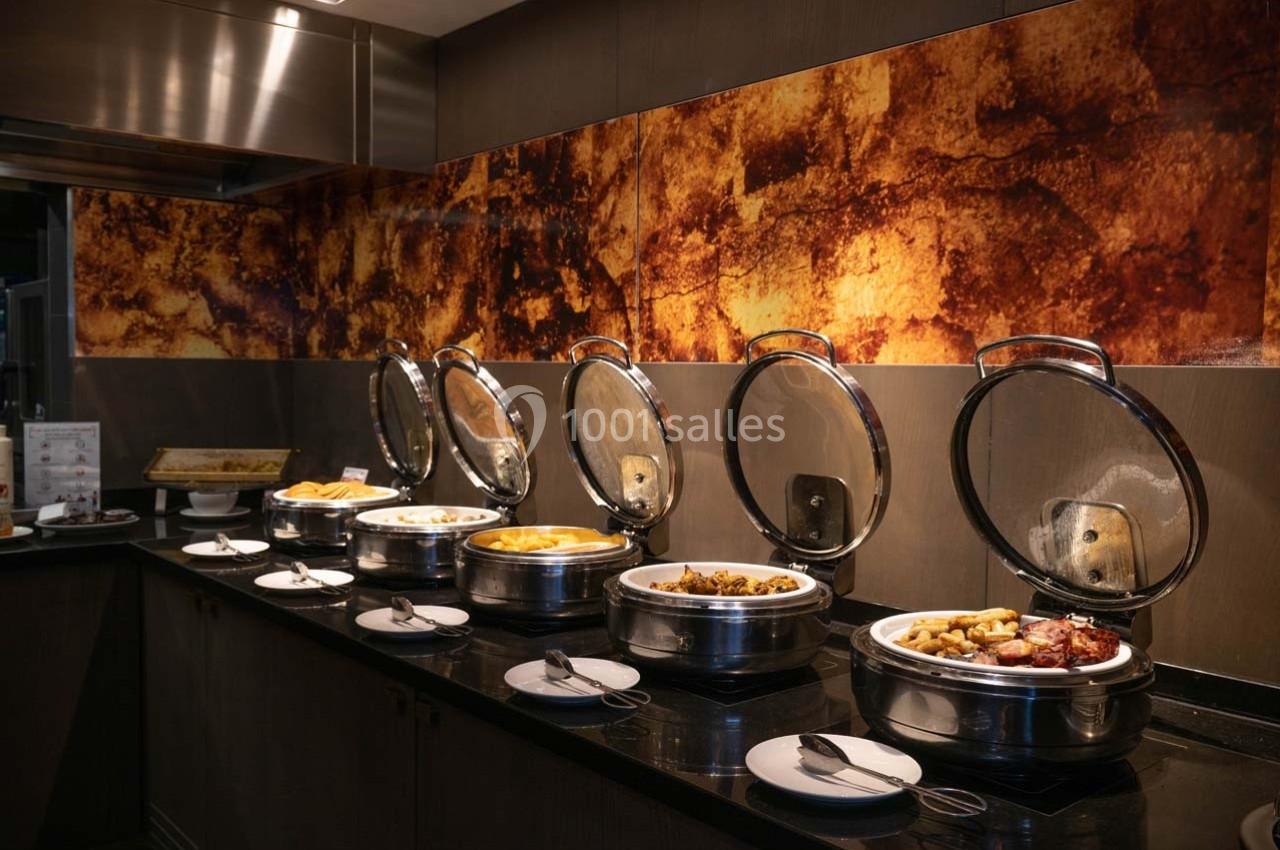 Buffet chaud avec plusieurs plats disposés dans des récipients en inox sur un comptoir sombre, décor mural texturé.