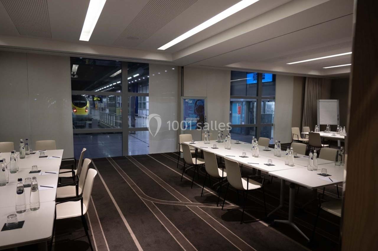 Salle de réunion moderne avec tables disposées en U, chaises blanches et vue sur une gare à travers de grandes baies vitrées.