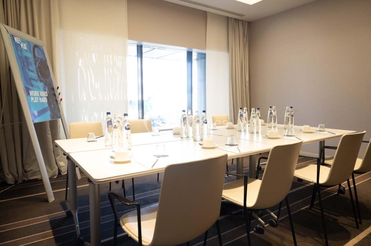 Salle de réunion lumineuse avec une table rectangulaire, des chaises, des bouteilles d'eau et un panneau en arrière-plan.