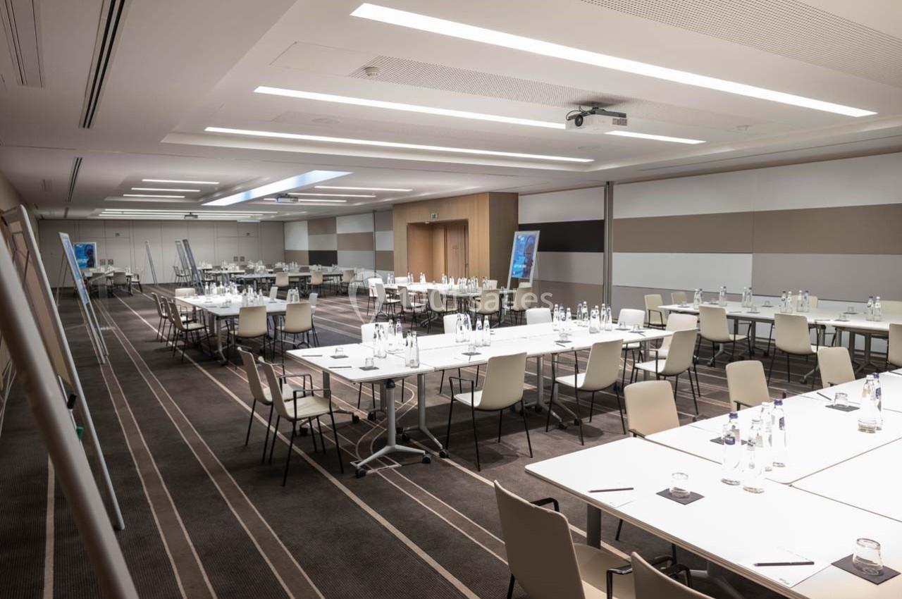 Salle de réunion moderne avec tables disposées en U, chaises alignées, bouteilles d'eau et écrans de présentation.