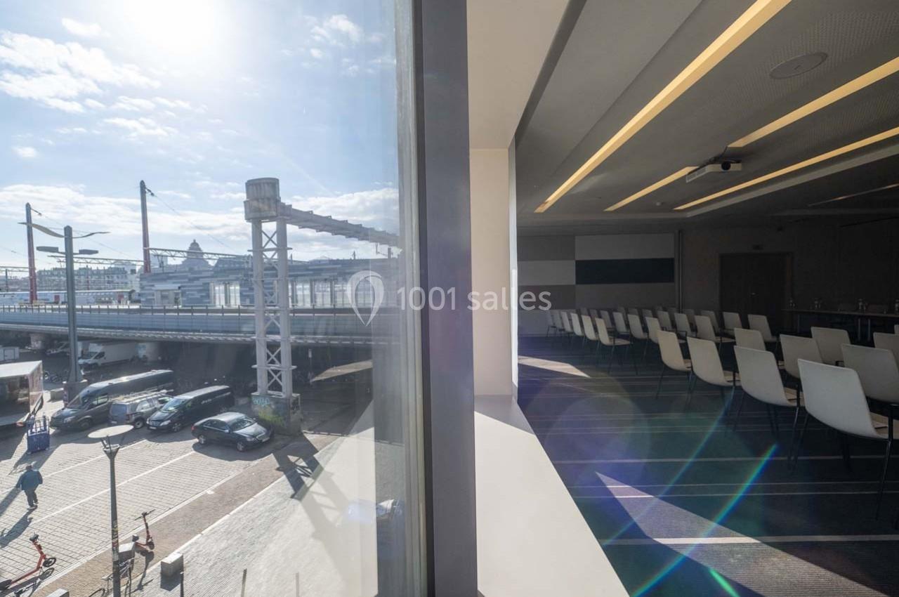 Vue depuis une salle de réunion vide avec des chaises blanches, donnant sur un quai et des structures industrielles…