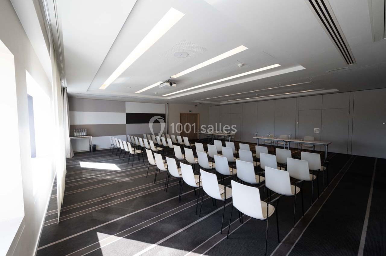 Salle de conférence lumineuse avec des rangées de chaises blanches alignées face à une table et un écran.