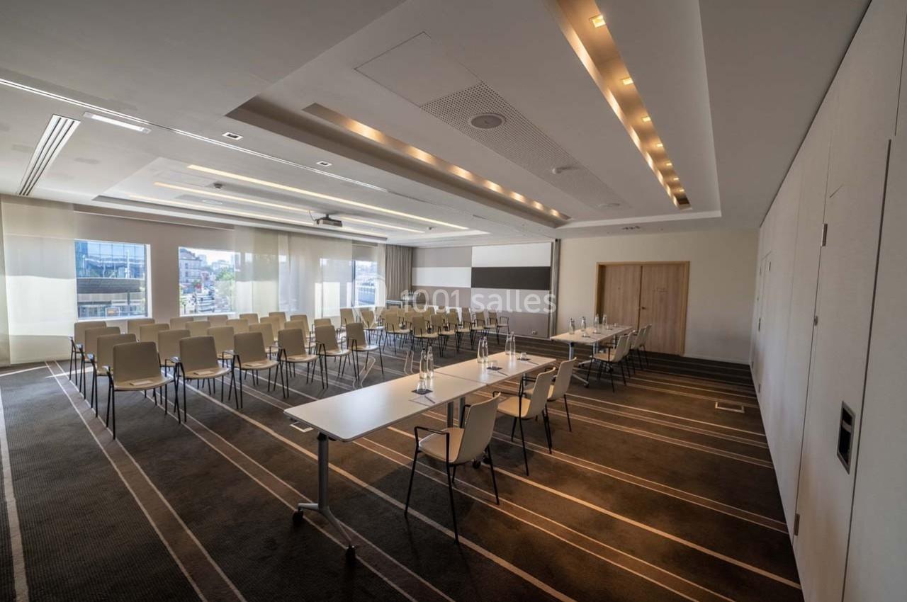 Salle de conférence lumineuse avec chaises alignées, tables équipées de verres et stylos, et grandes fenêtres vitrées.